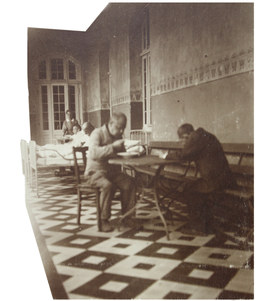 Schwarzweiß Foto zweier Männer, die an einem Tisch sitzen, im Hintergrund ein Krankenbett.